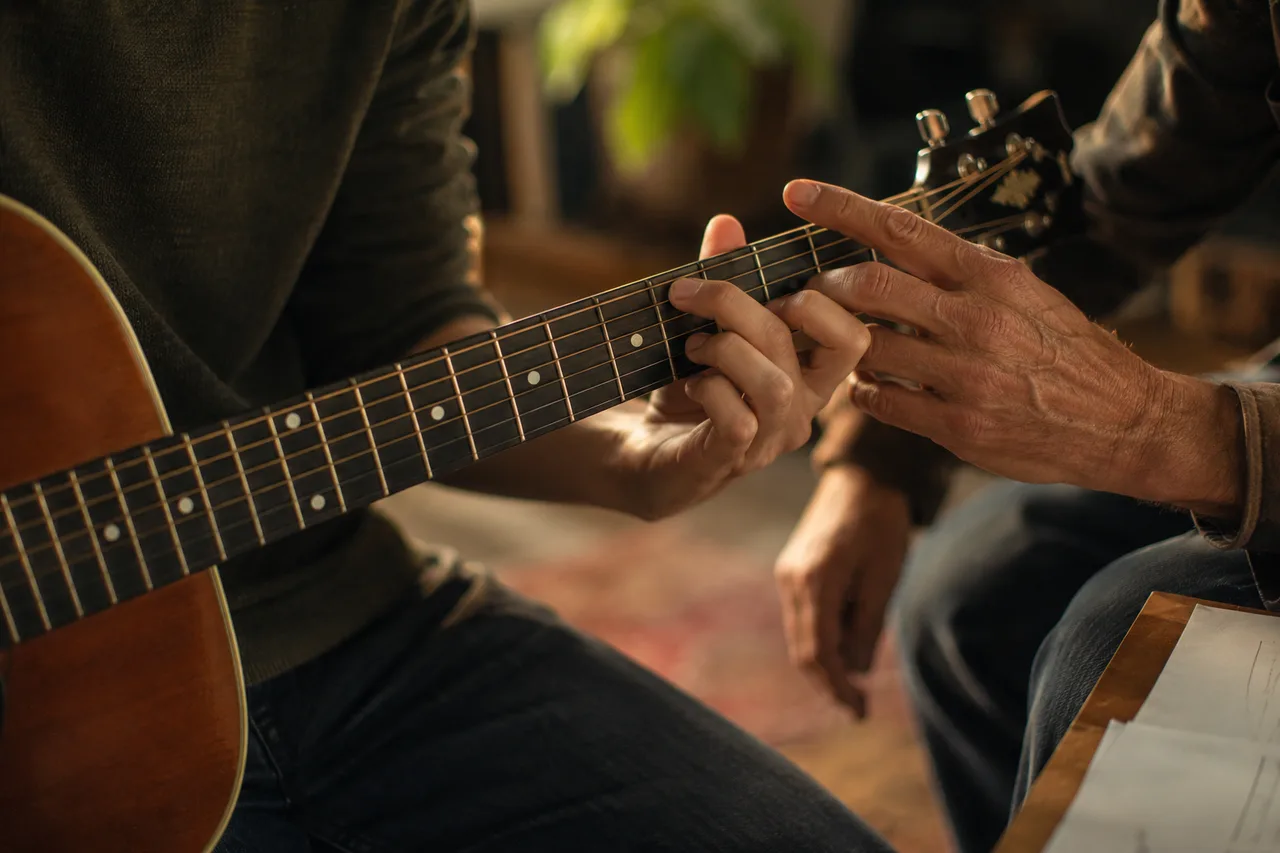 A student's hands forming a guitar chord while a teacher's hand gently adjusts one finger